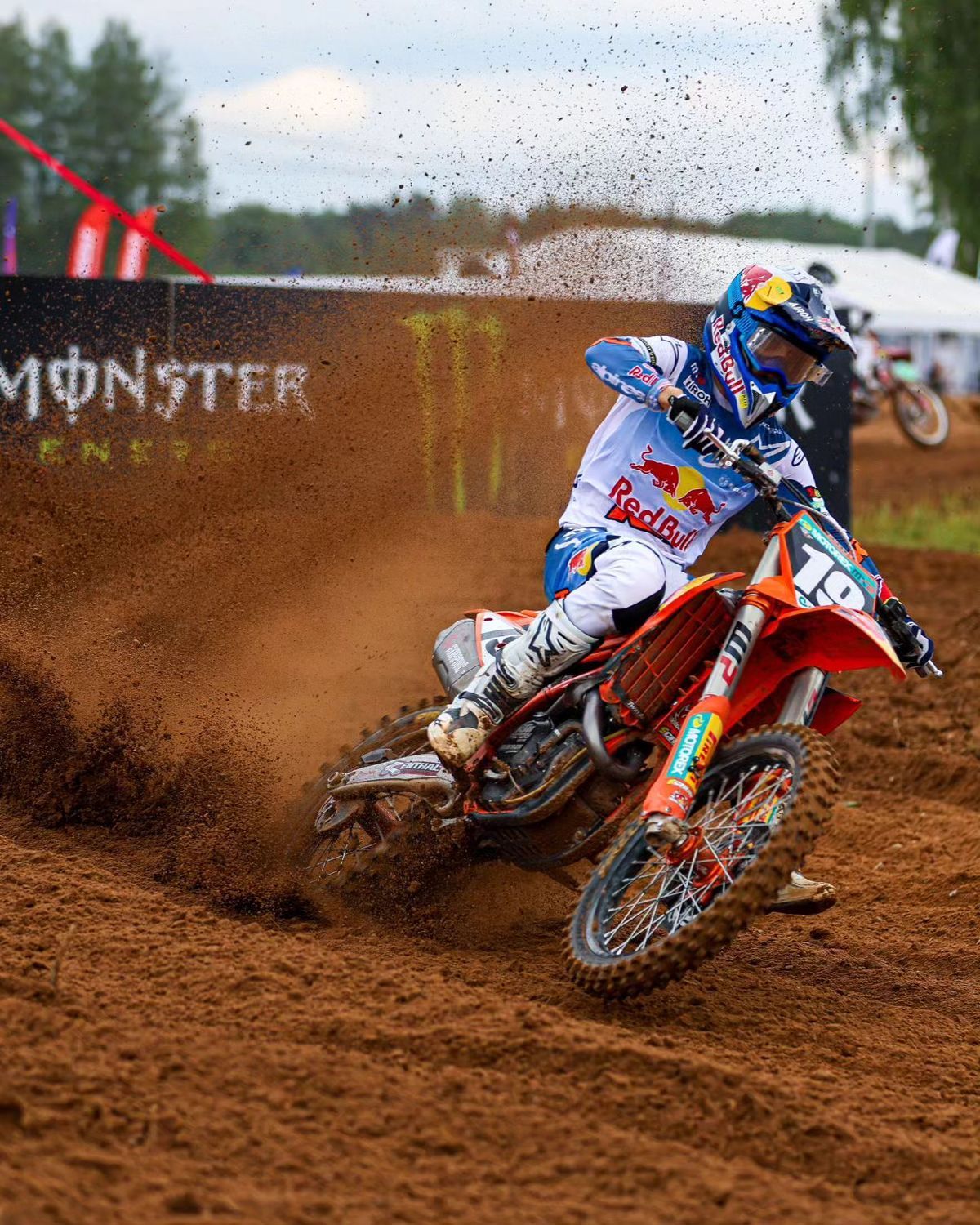 Motocross rider on an orange bike, kicking up dirt, with Monster Energy branding.
