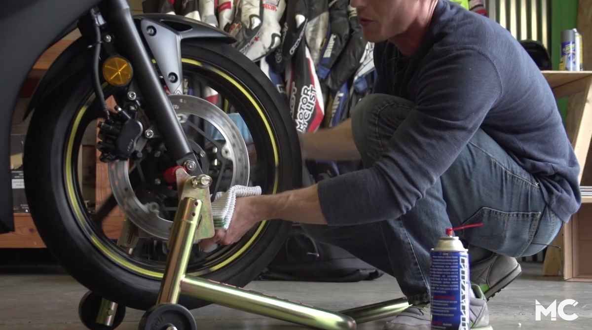 Man cleaning a motorcycle brake disc with a cloth, standing in a garage, using a spray can.