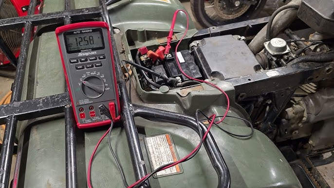 A red multimeter on a green ATV measures 12.5 volts at the battery. Wires connect the meter to the battery terminals.
