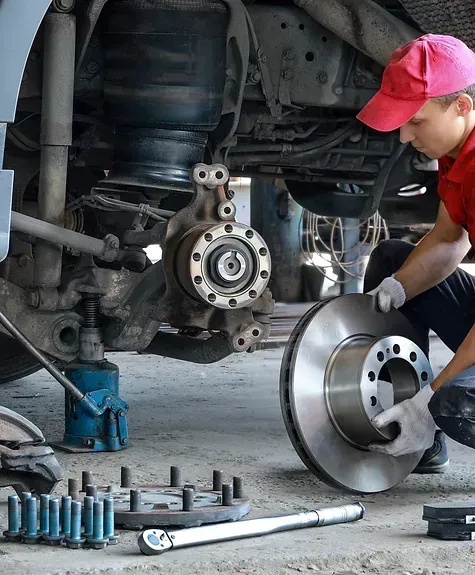 Mechanic installing brake rotor on a vehicle. Wears red shirt, cap, and gloves, working in a garage.