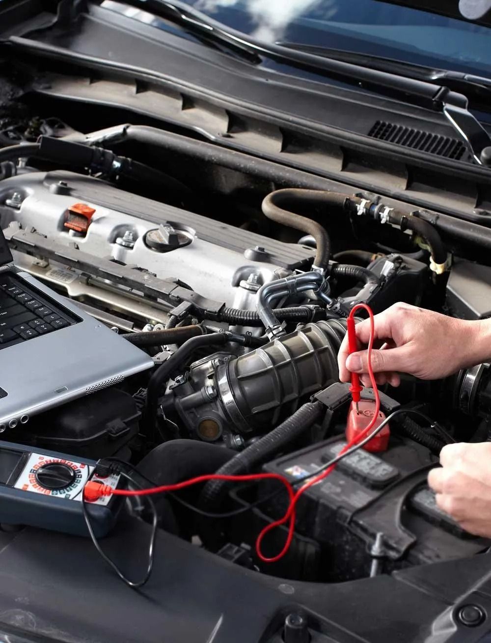 Car engine being tested with multimeter and laptop in an open hood.