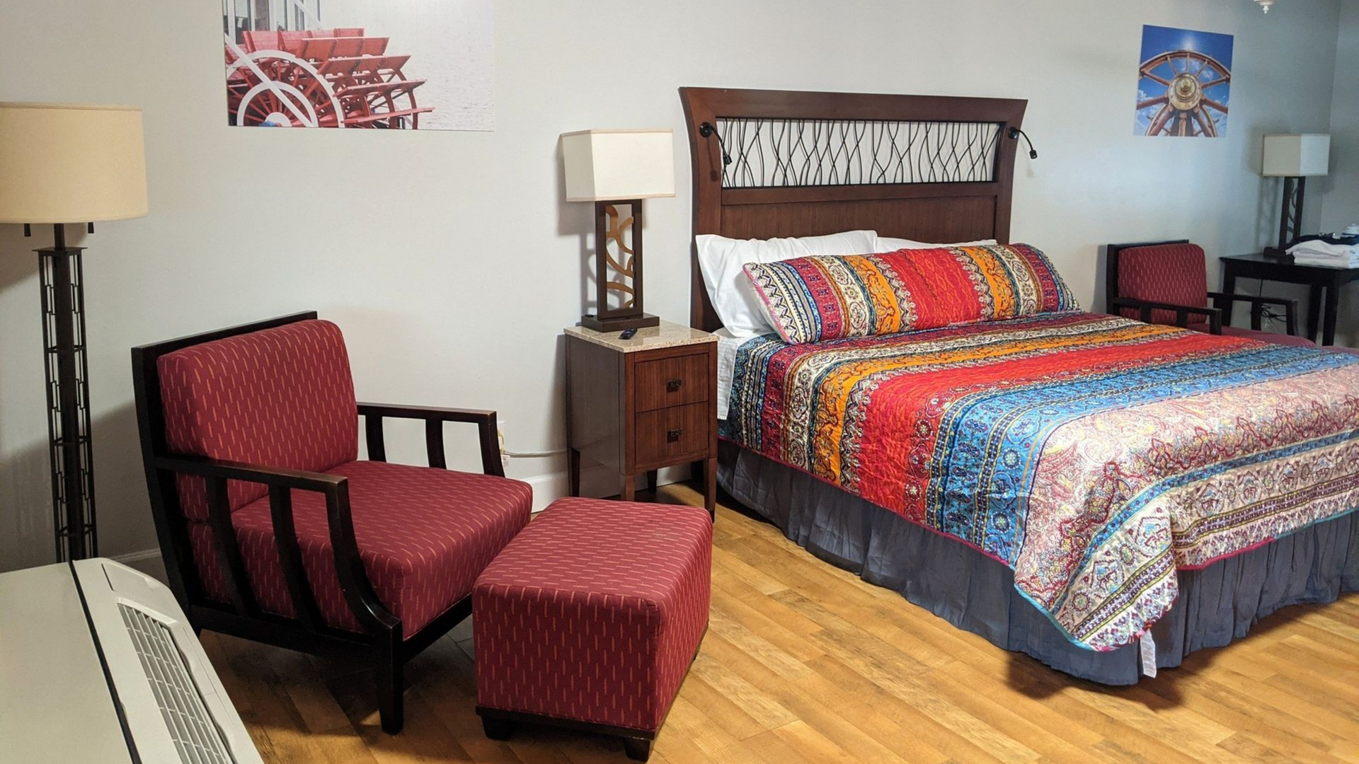 Hotel room with a bed, red armchair and ottoman, two lamps, and wall art on hardwood floors
