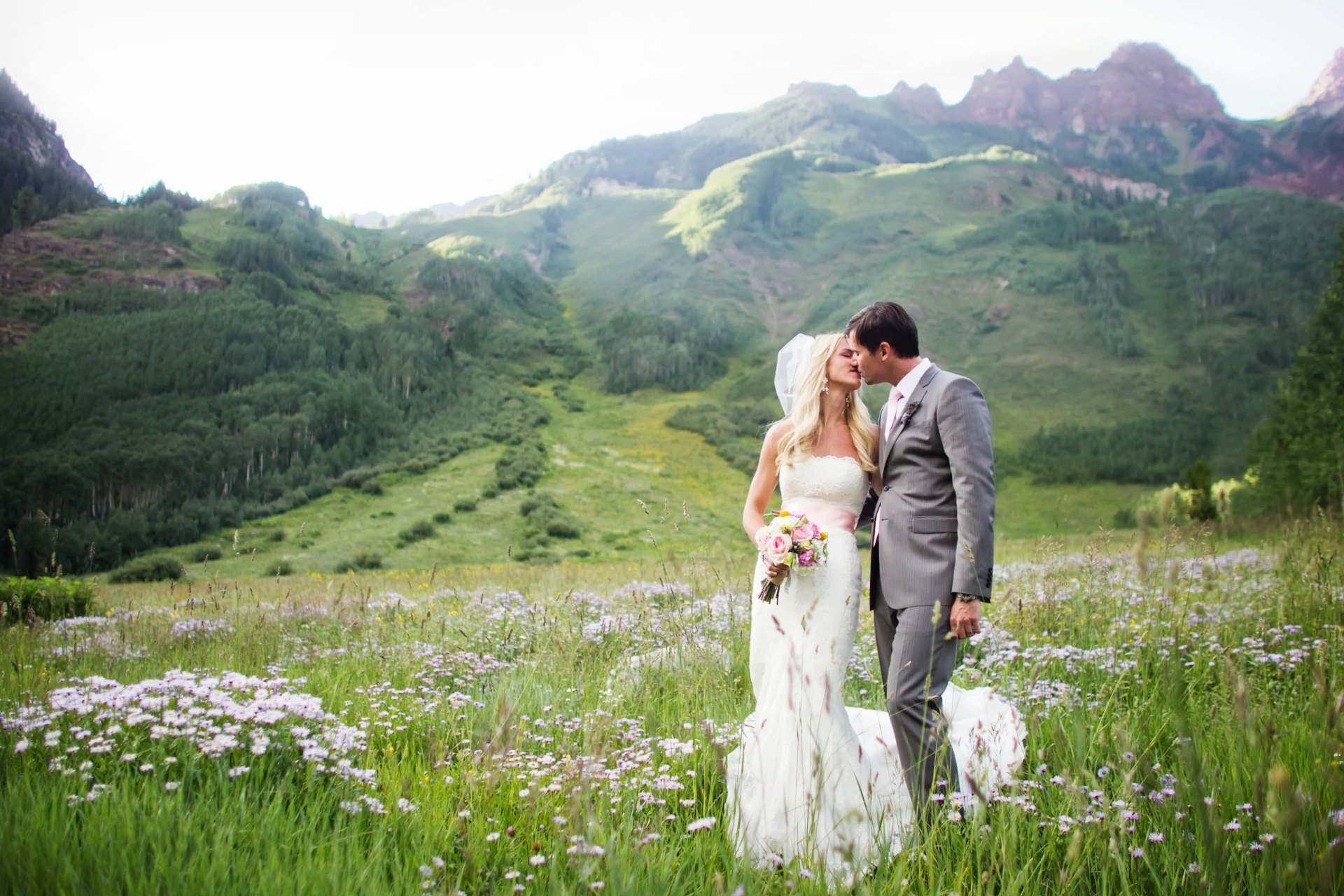 a bride and groom are kissing in a field with mountains in the background .