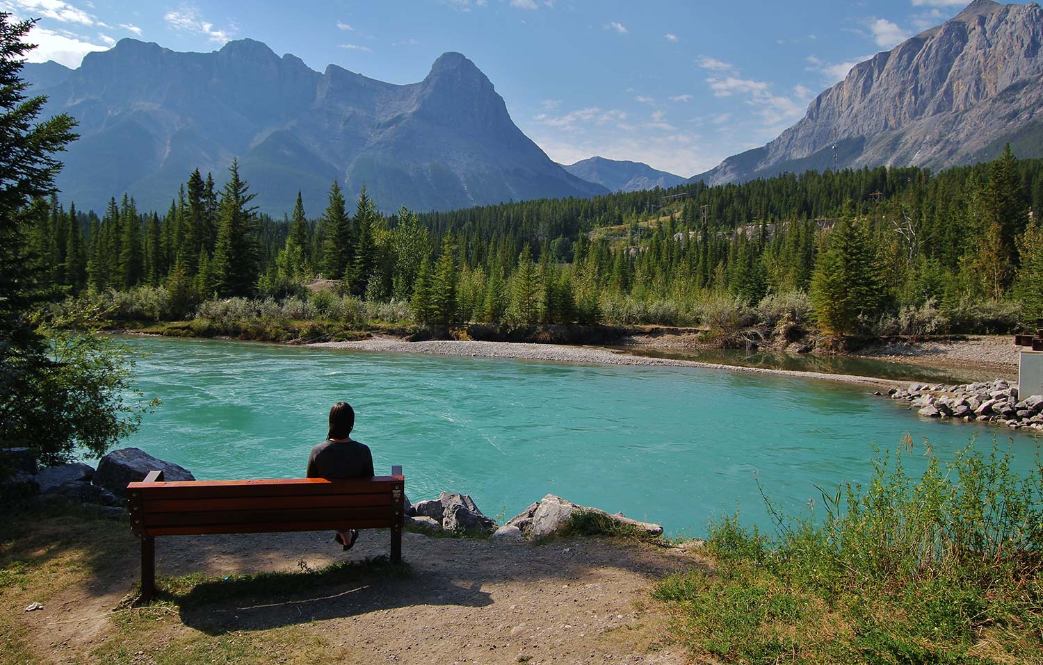 A person sits on a bench overlooking a lake with mountains in the background