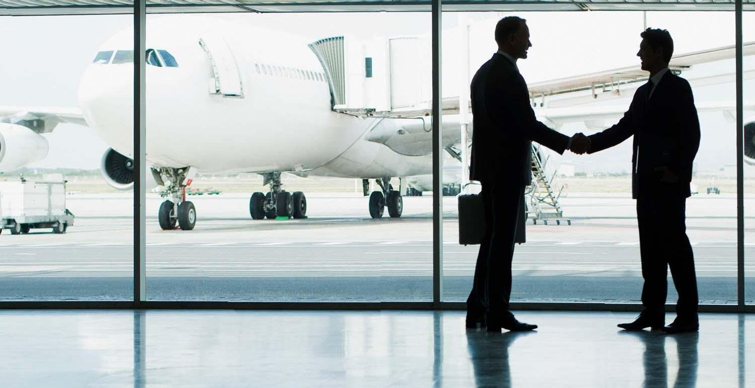 Two men are shaking hands in front of a window with an airplane in the background.