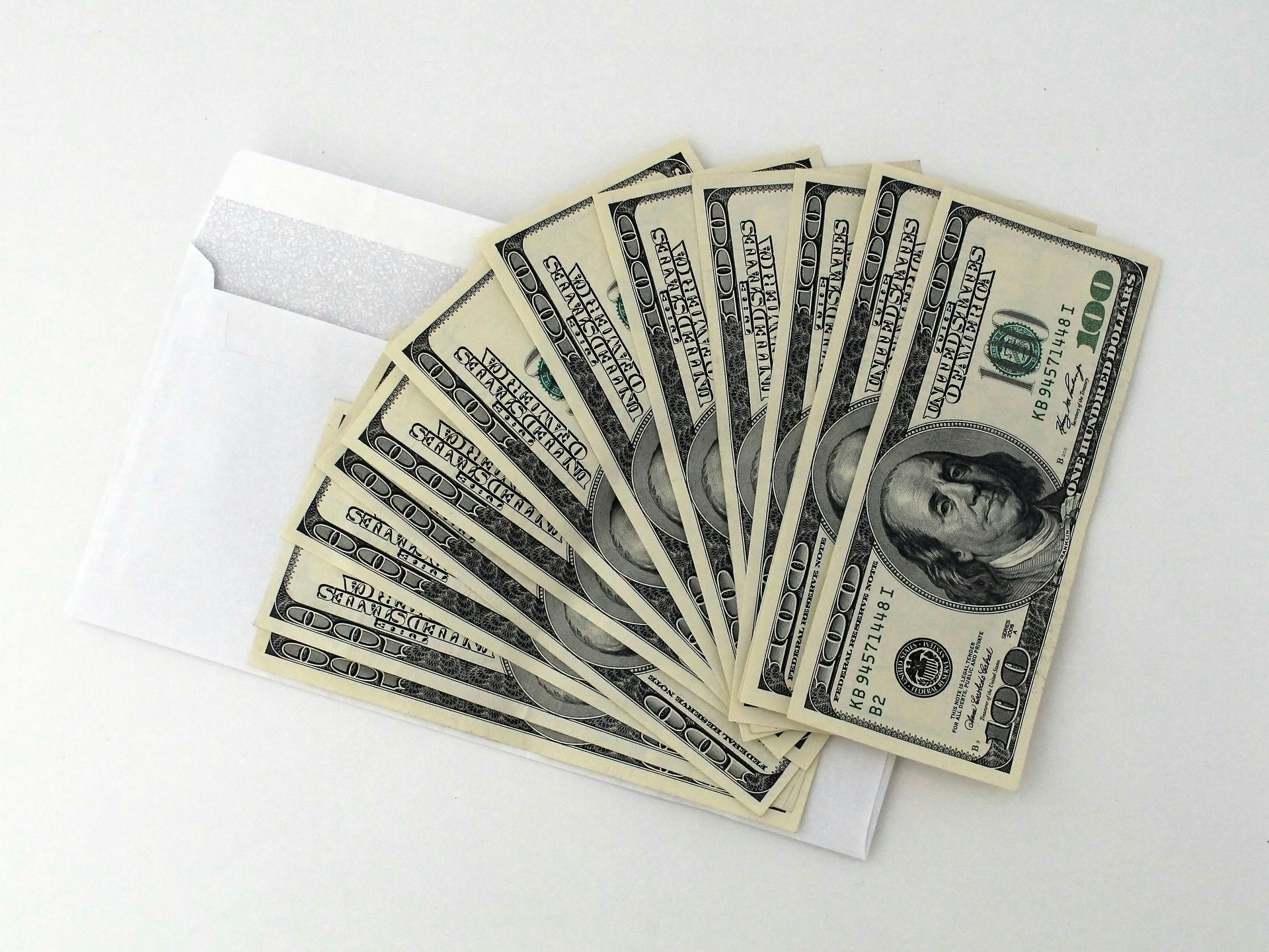 Fan of ten $100 bills extending from a white envelope on a white surface.