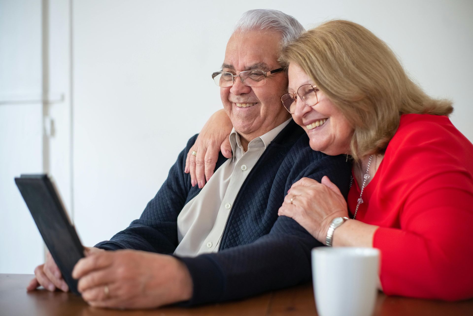 Smiling couple looking at a tablet together, woman's arm around man. They are inside, near a white wall.