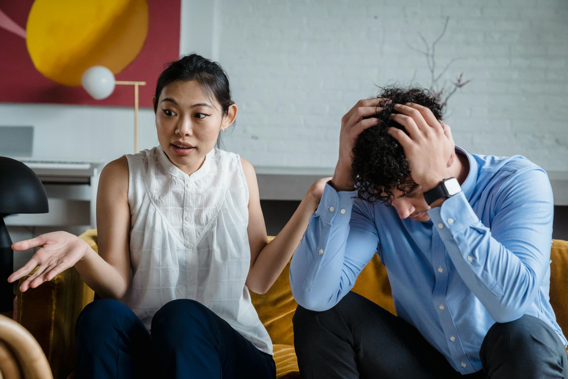 Woman gesturing, man with head in hands, both on a couch, appearing upset.