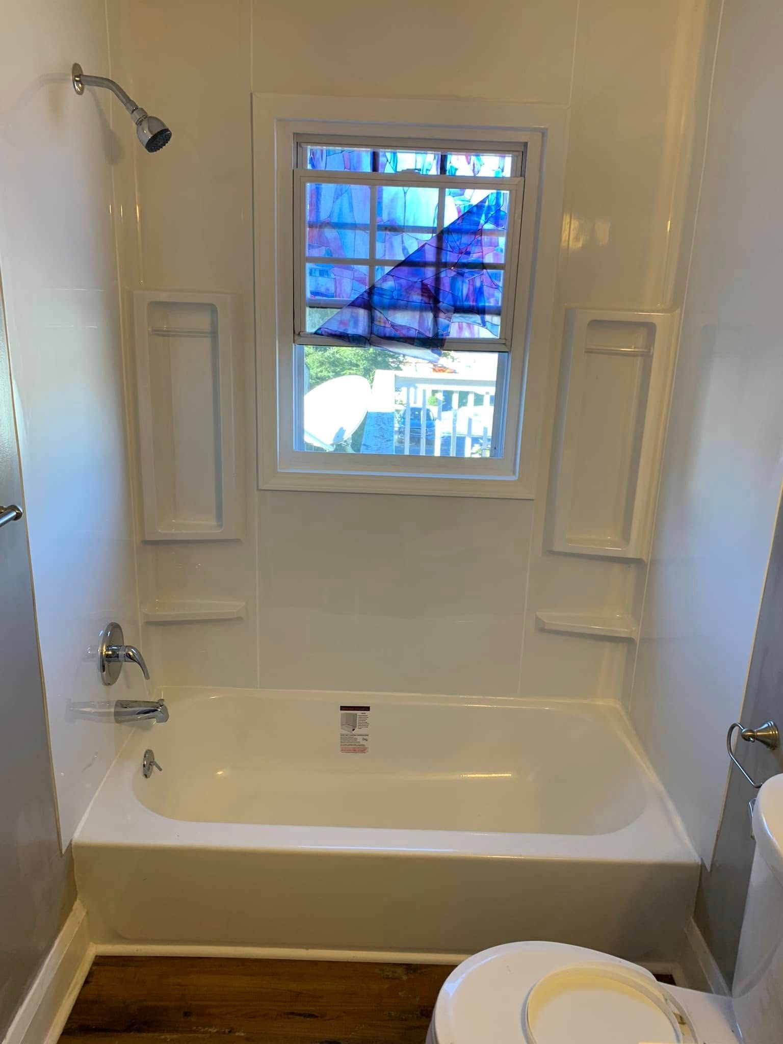 A white bathtub and shower surround with a window above, set against neutral walls in a bathroom with wood-look flooring.