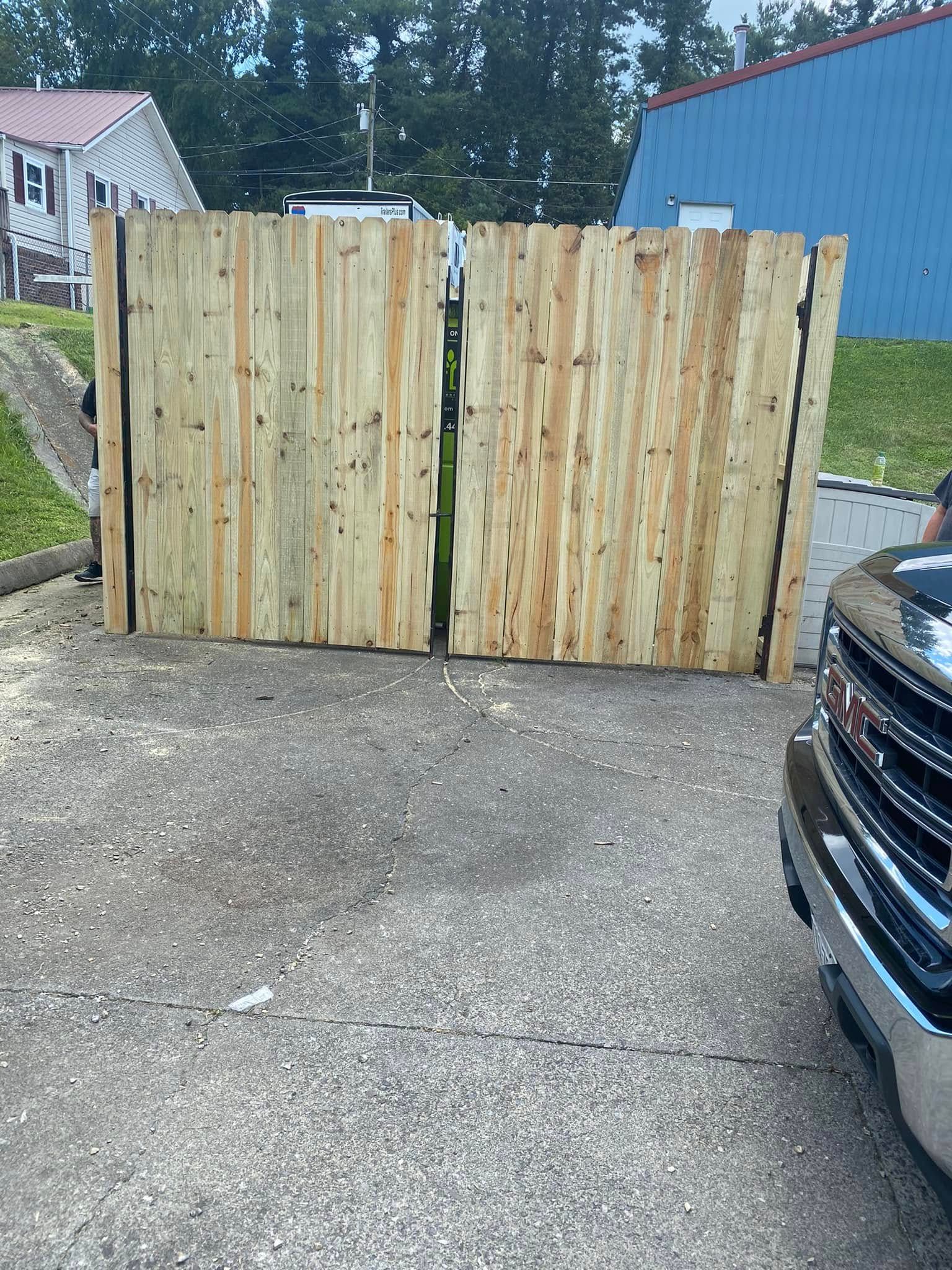 A pair of light-colored, vertical wooden fence gates installed on a concrete driveway, next to a black vehicle.