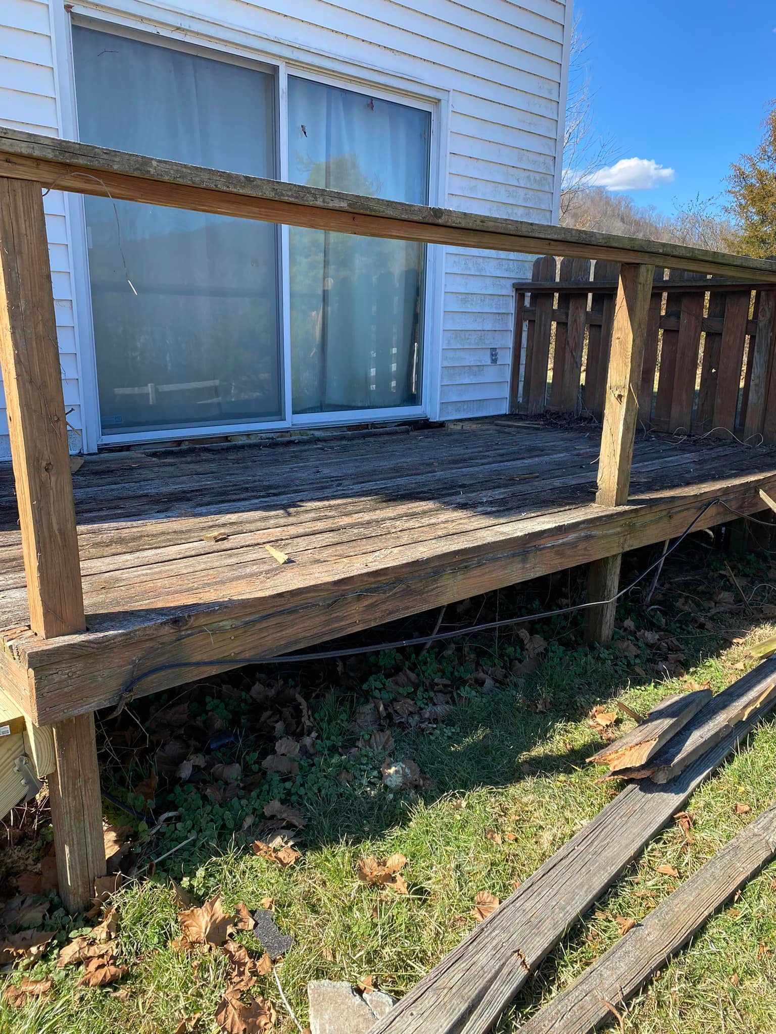 A weathered wooden deck with a railing sits outside a house with white siding and a sliding glass door.