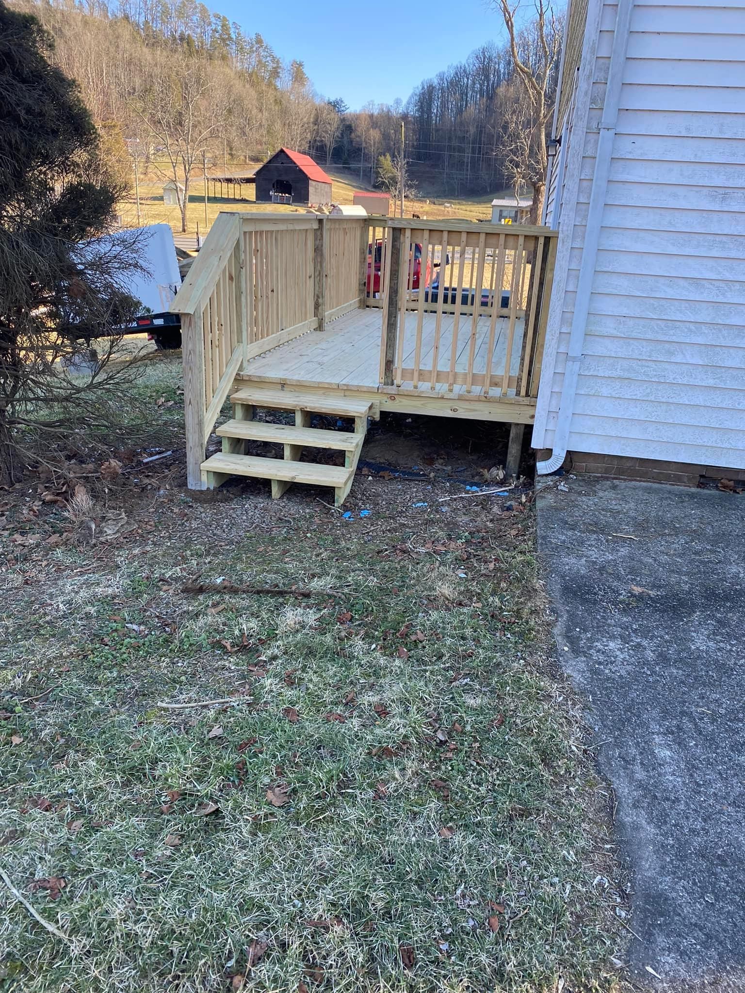 A newly built wooden deck with stairs attached to the side of a white house on a grassy lot with a barn in the background.