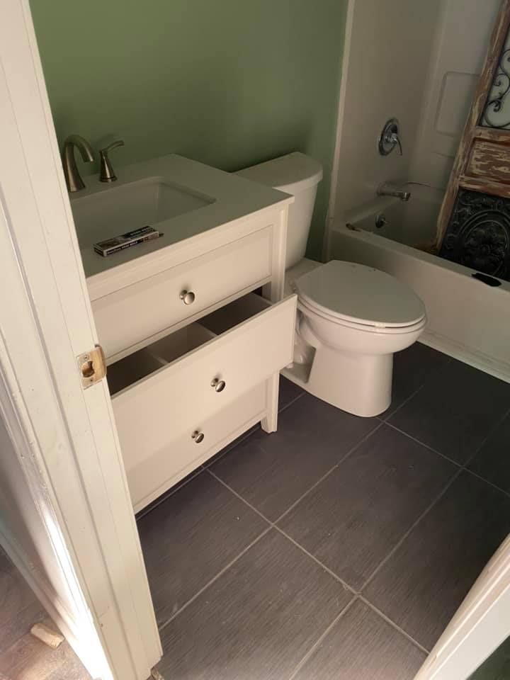 A bathroom interior featuring a white vanity with open drawers, a white toilet, and gray tile flooring.