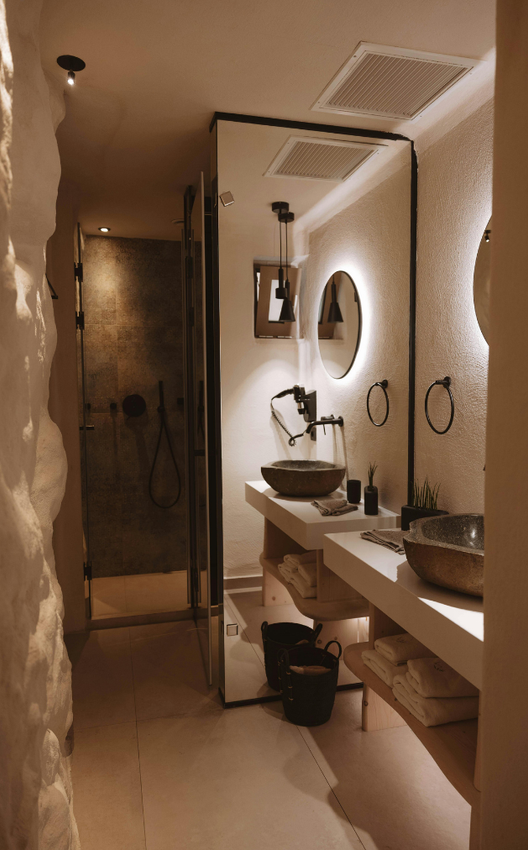 Contact Us A modern, dimly lit bathroom with dual stone sinks, a large mirror, and a walk-in shower with textured, light-toned walls.