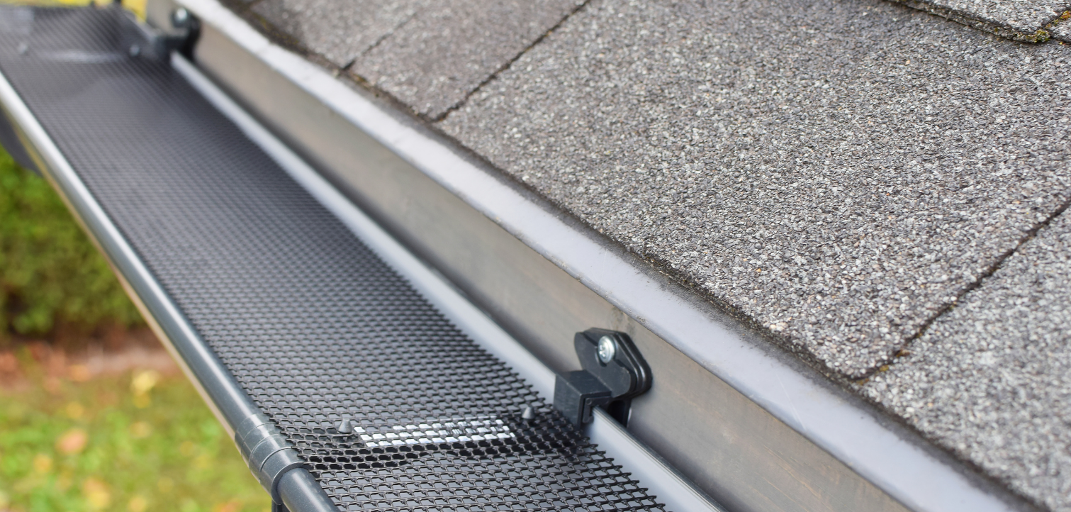 Contact Us A black mesh gutter guard installed on a rain gutter beneath the edge of a shingled roof.