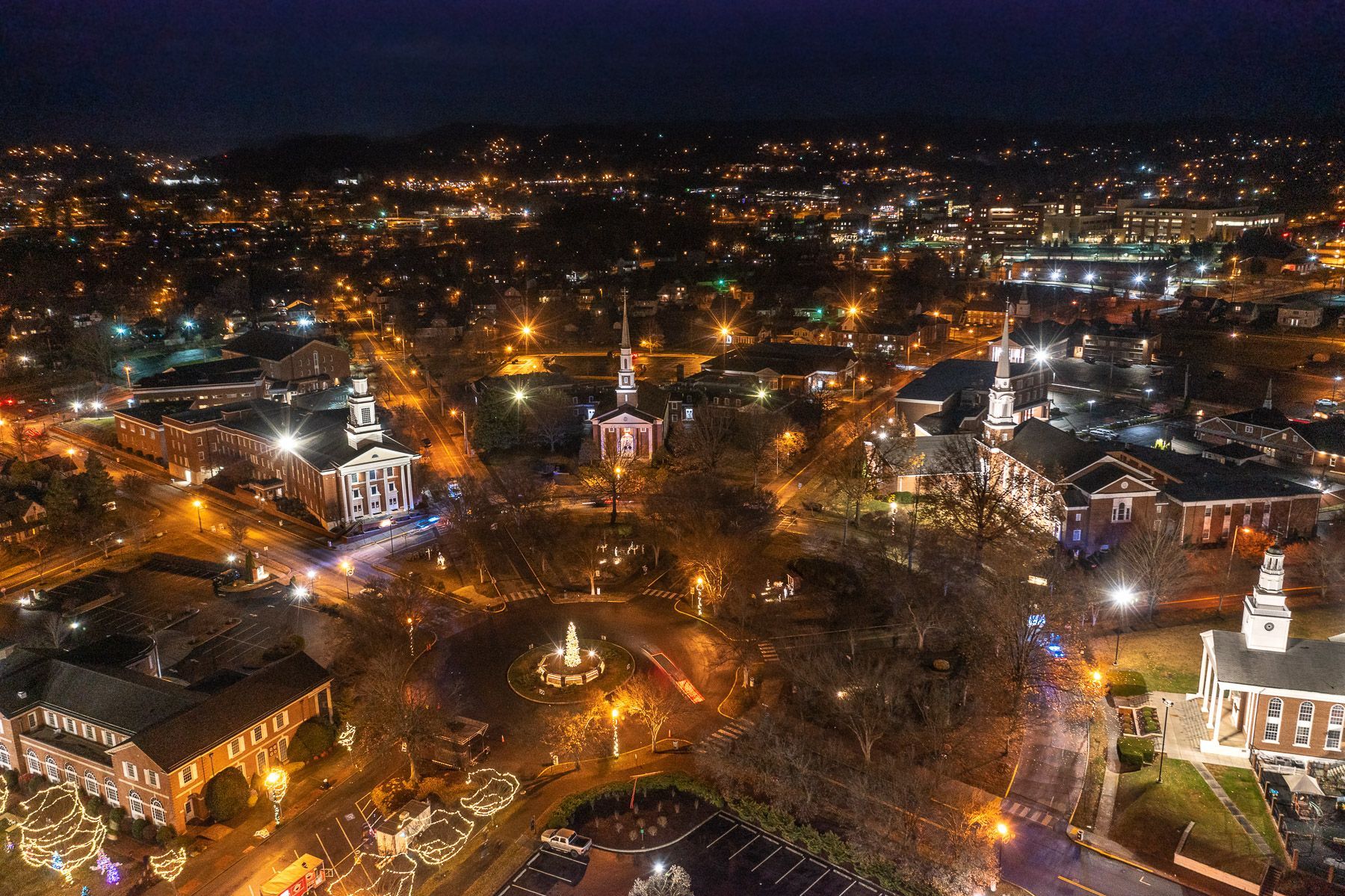 Aerial night view of a glowing town square with several churches, street lights, and a central fountain in Kingsport