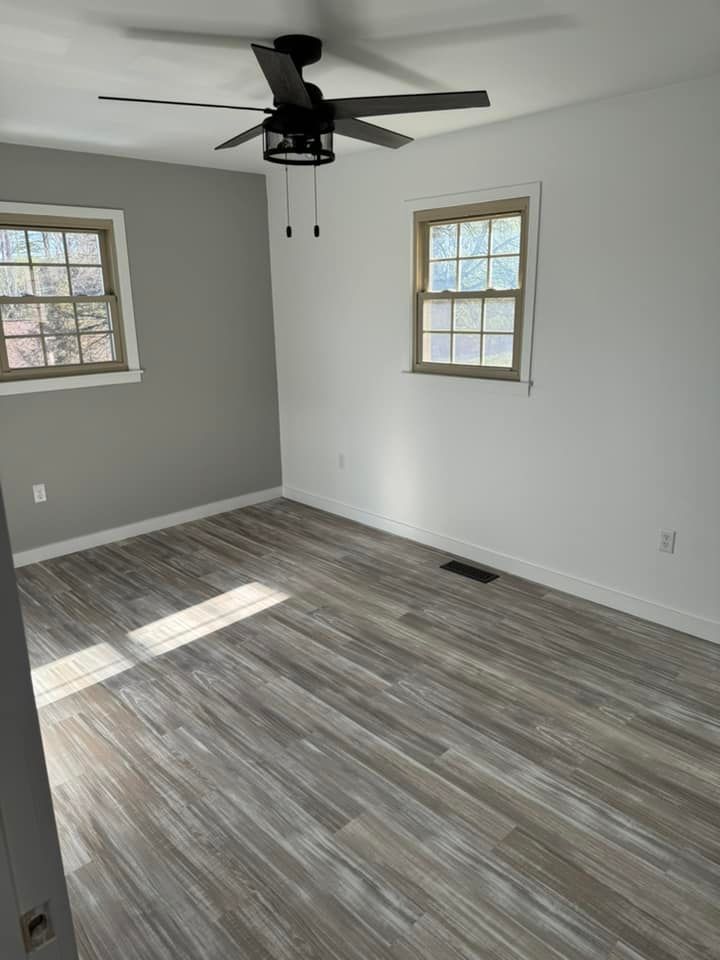 An empty room with light gray walls, wood-look flooring, two windows, and a dark ceiling fan.