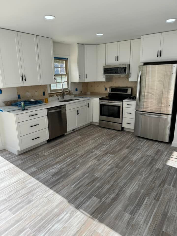 A modern kitchen with white cabinets, stainless steel appliances, and grey wood-look flooring.