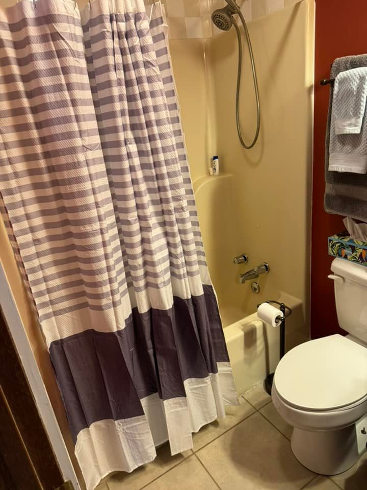 A bathroom featuring a shower with a gray and white striped curtain, a beige tub, and a toilet next to a red wall.