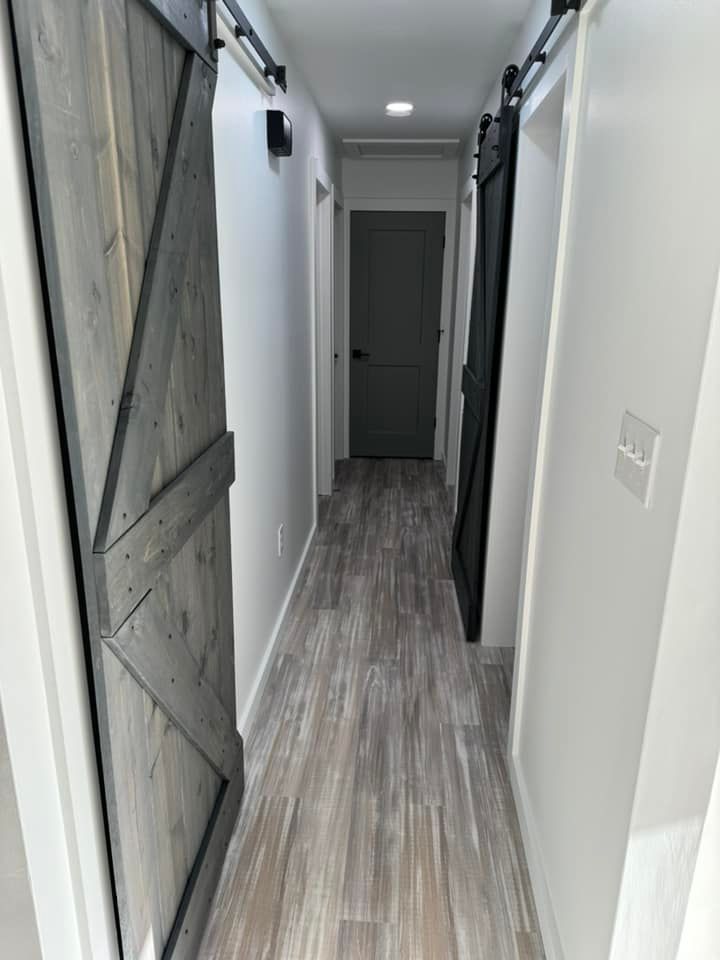 A narrow hallway with grey wood-look flooring, white walls, a light-grey barn door, and a dark-grey door at the end.