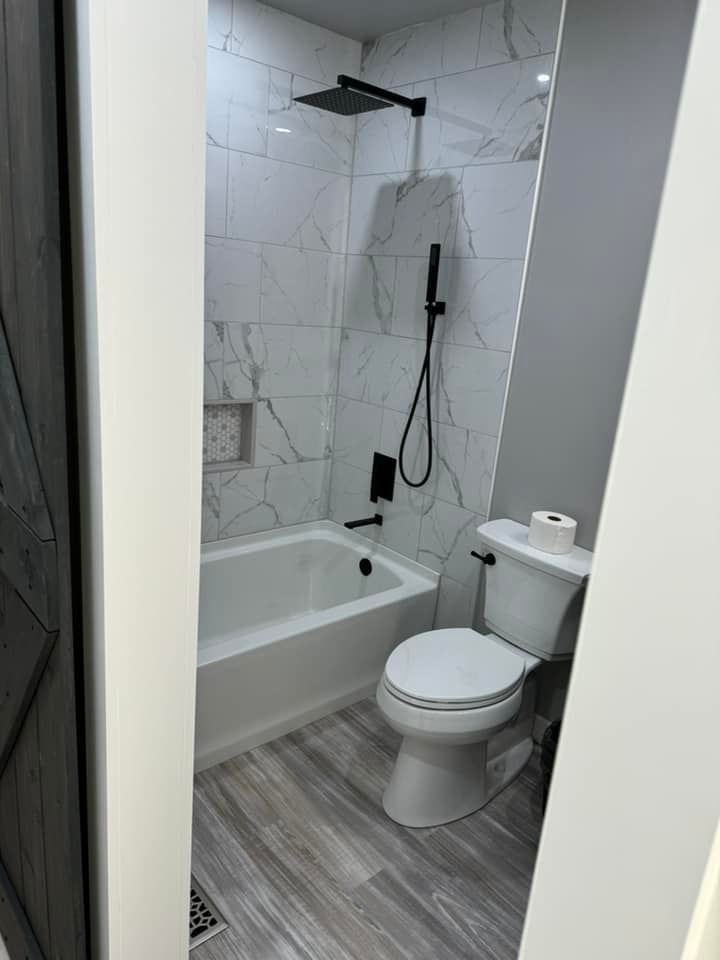 A small modern bathroom featuring a white bathtub with white marble-patterned tile, black fixtures, and a white toilet.