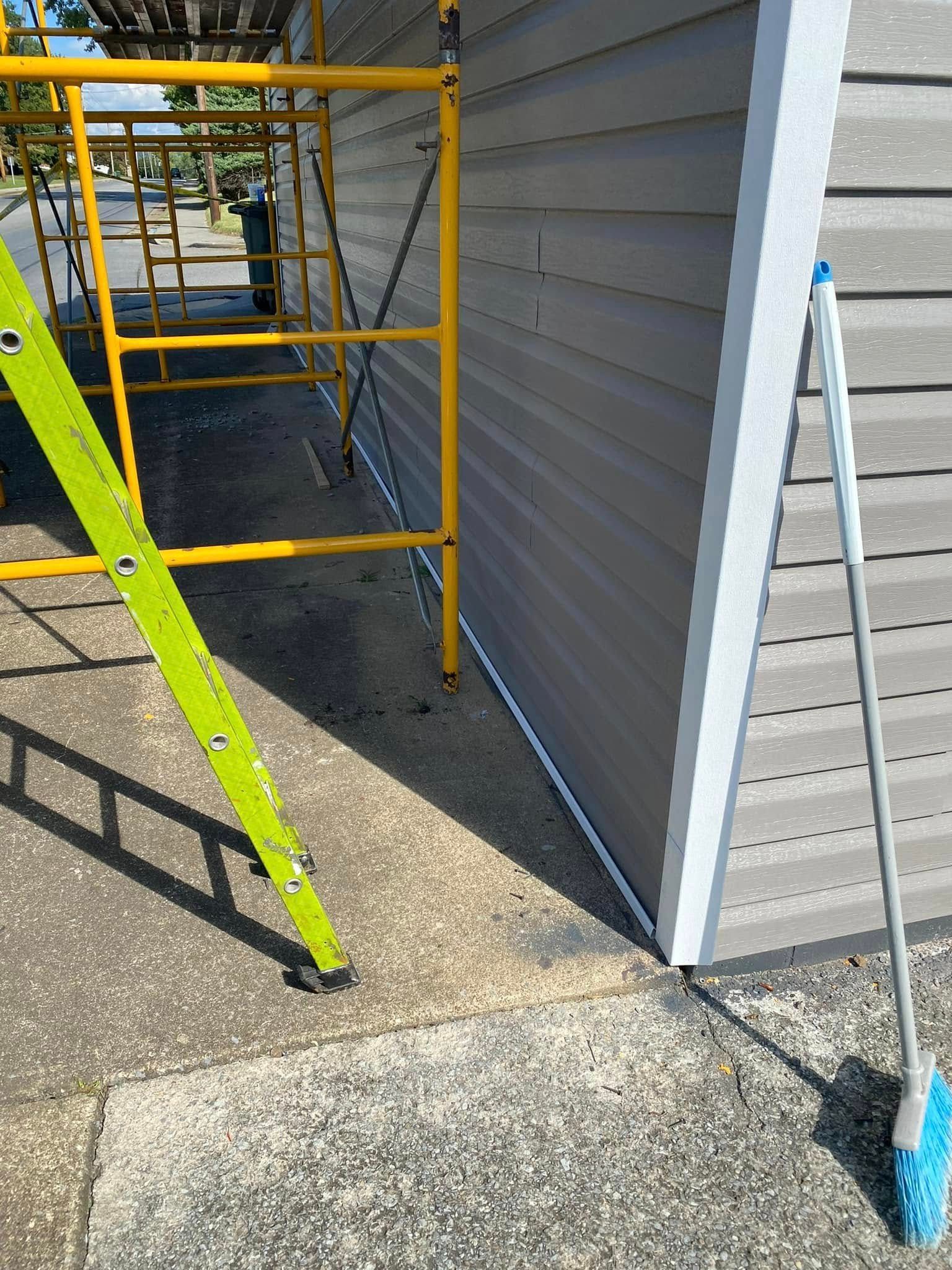 A yellow metal scaffold stands next to a grey-sided building, with a green ladder and a blue-headed mop leaning nearby.