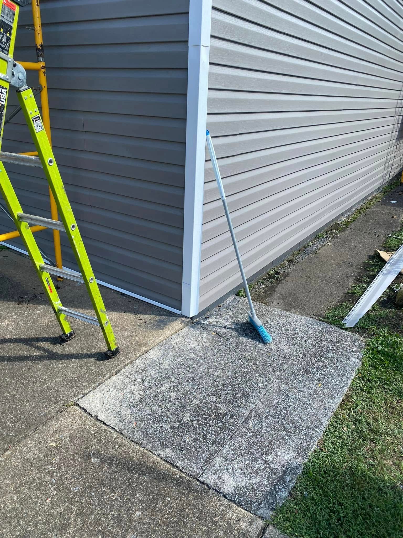 A yellow ladder stands beside the grey-sided corner of a house near a concrete walkway and a blue cleaning tool.
