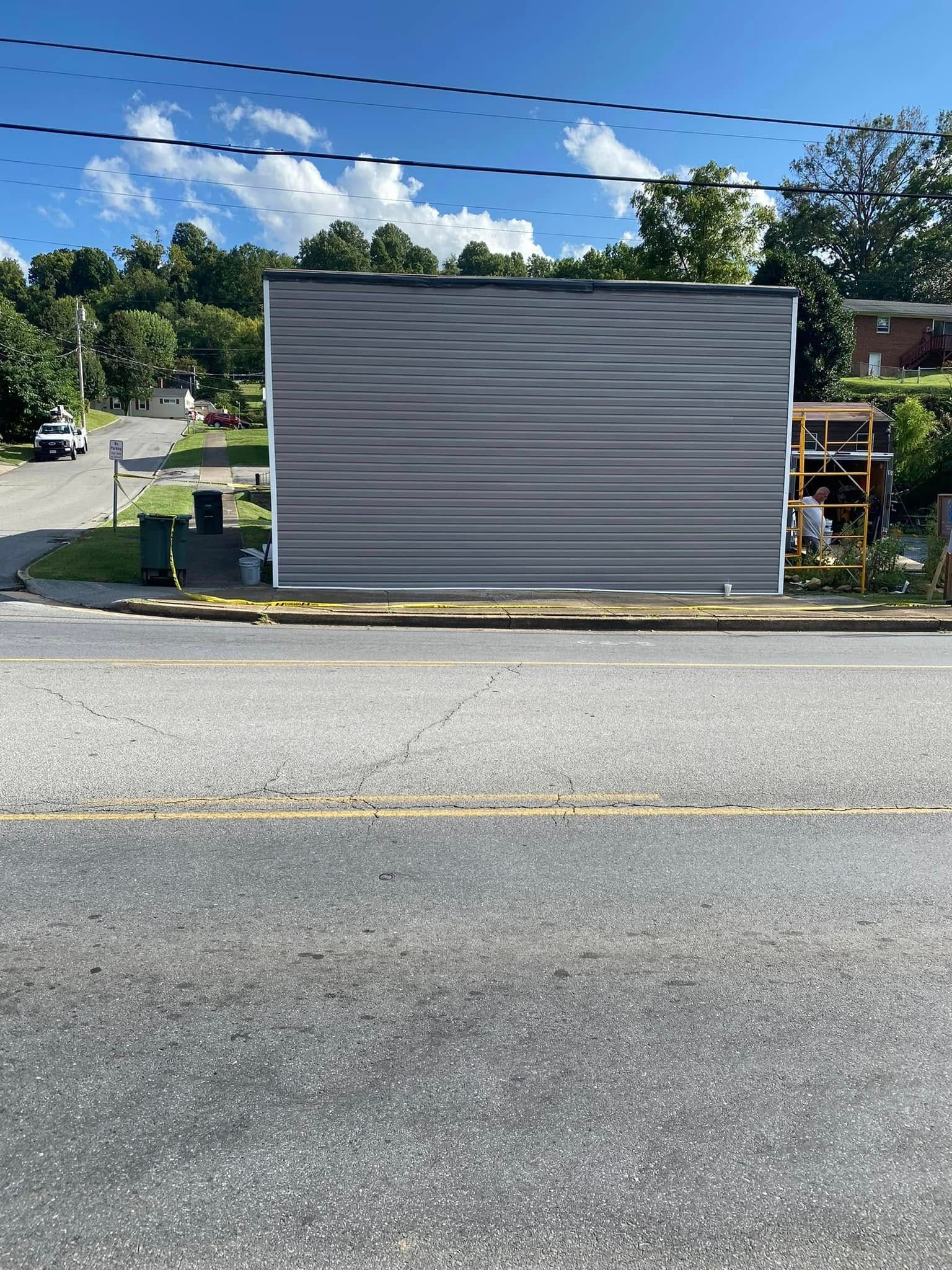 A gray, horizontal-sided building wall faces a paved road, with a partially visible wooden structure and trees behind it.