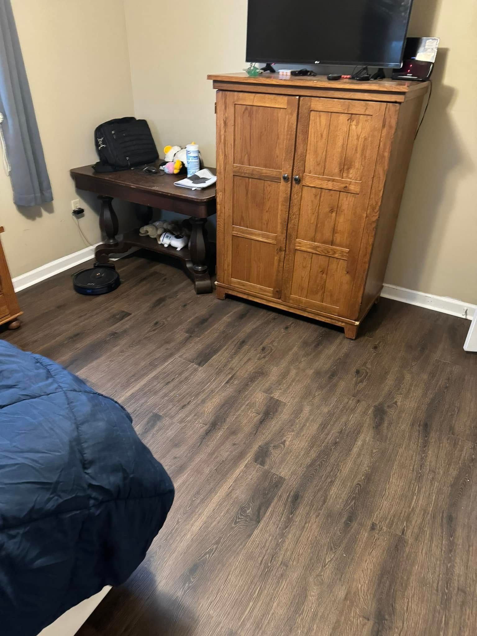 A bedroom featuring dark wood-look floors, a wooden armoire with a TV on top, a small writing desk, and a blue bedspread.