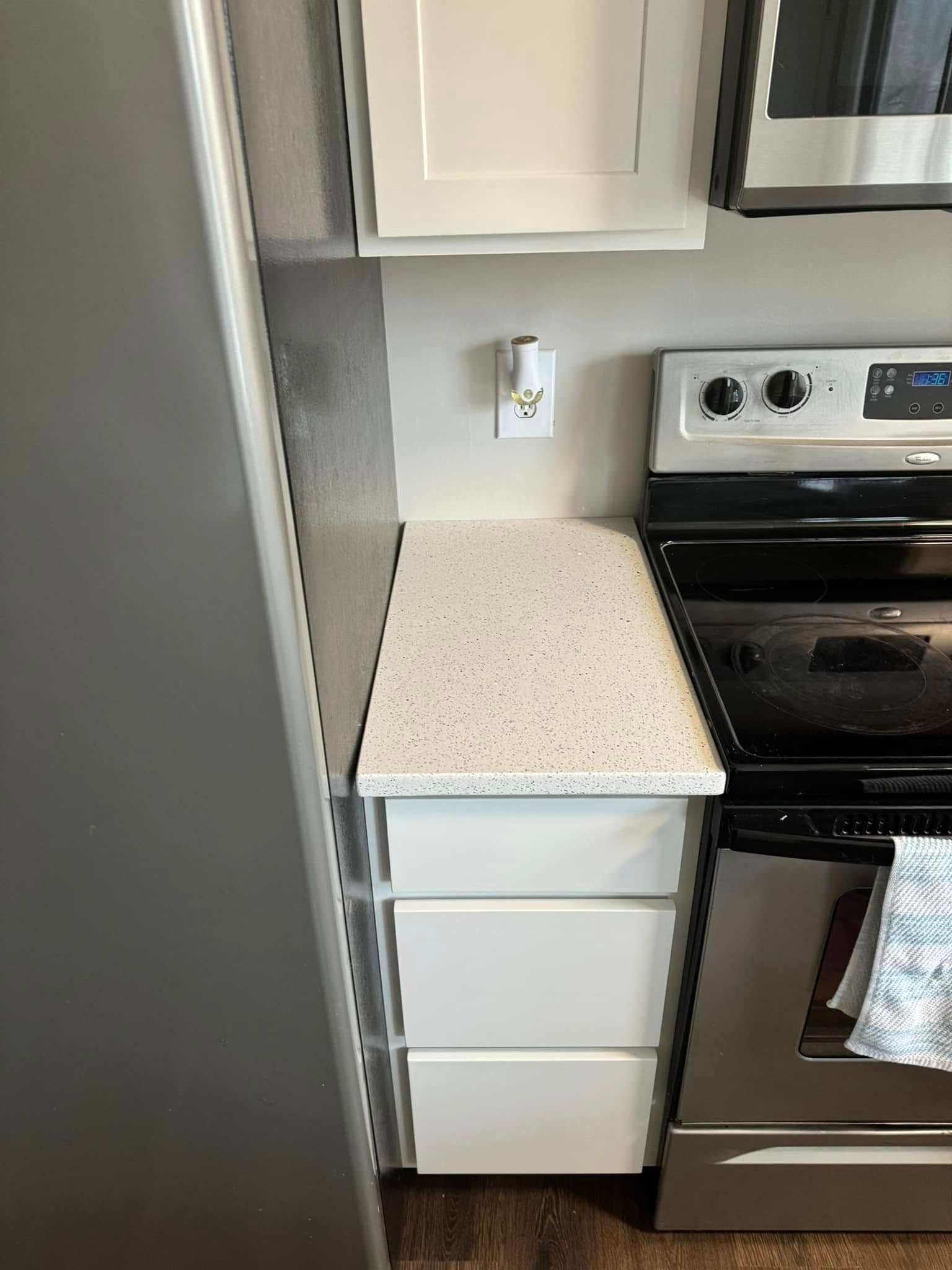 A white three-drawer cabinet sits between a gray wall and a stainless steel oven in a kitchen.