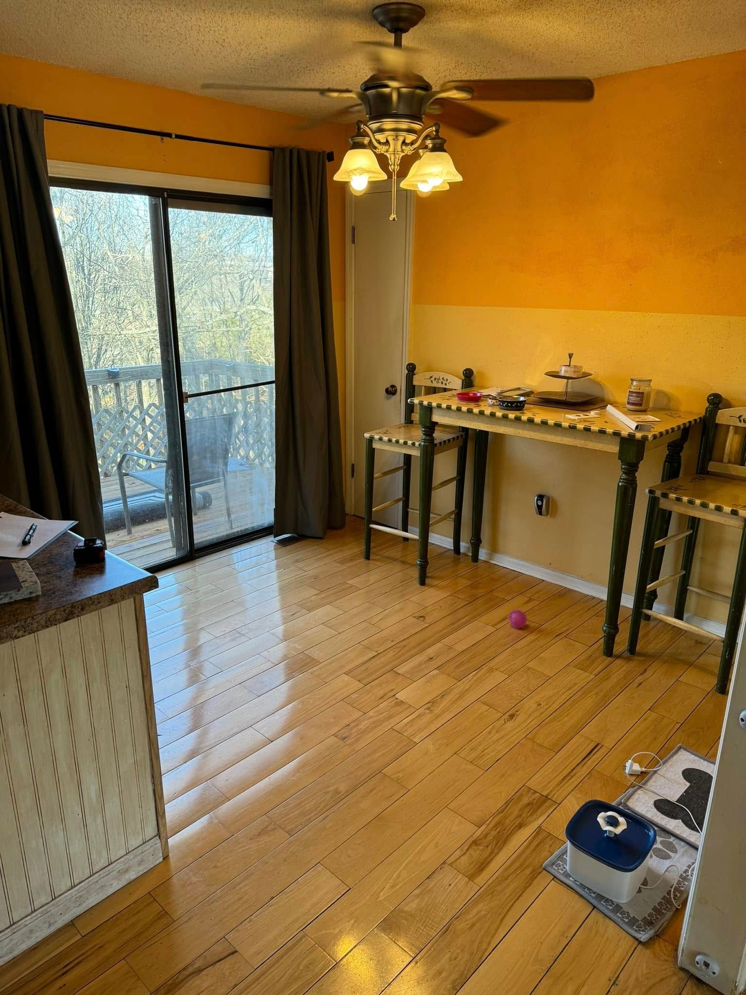 A dining area with wood floors, a high-top table with two stools, an orange wall, and a sliding glass door with curtains.