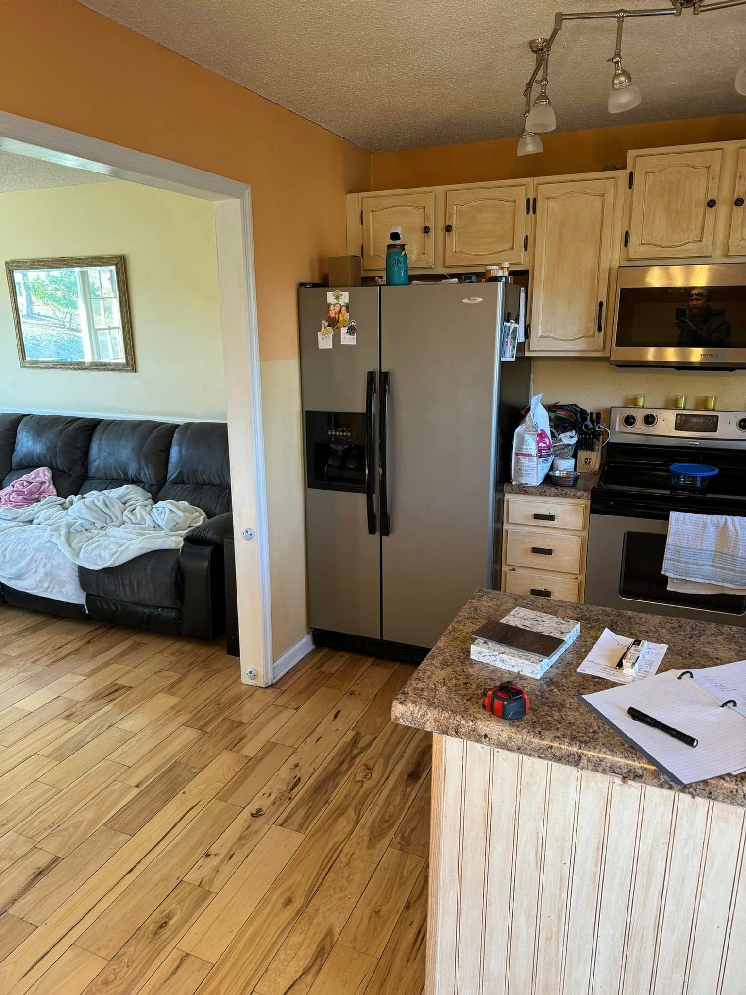 A view of a kitchen with light wood cabinets, stainless steel appliances, and an island, next to a living room sofa.