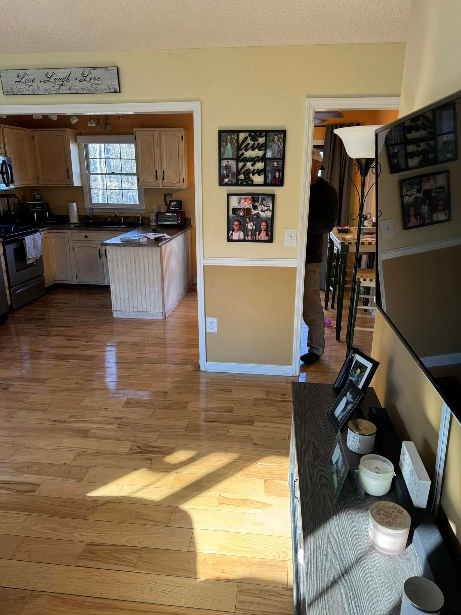 A bright, wood-floored kitchen and dining area seen from a living room, with family photos on the dividing wall.