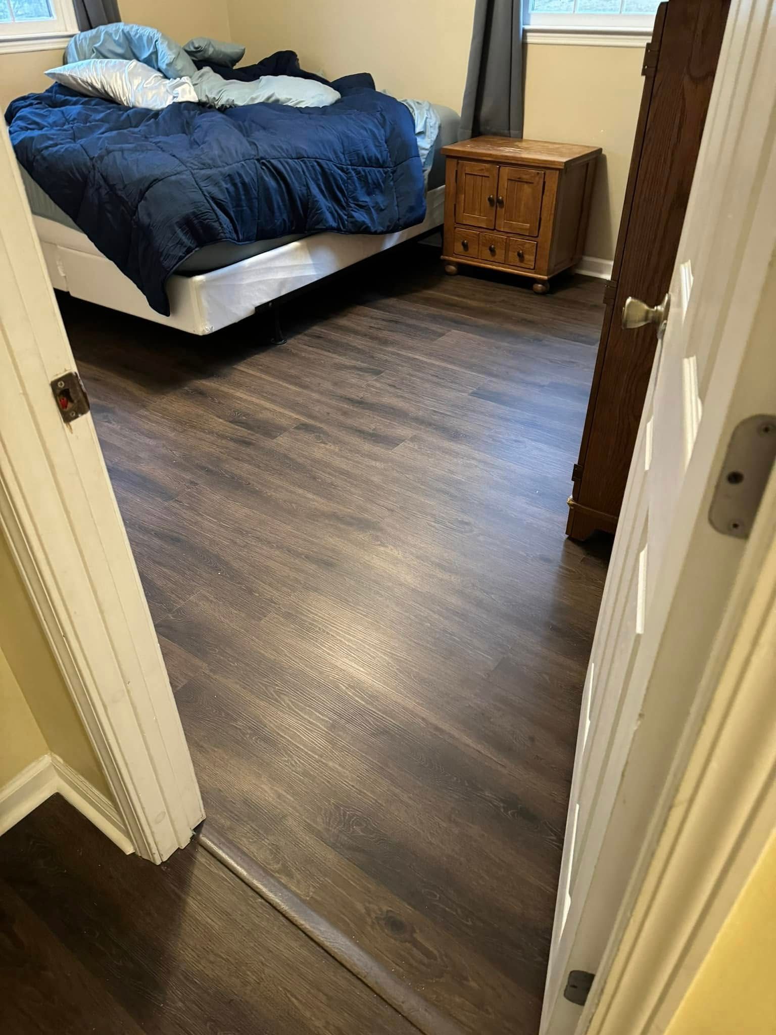 A bedroom view from a doorway, featuring dark wood-look laminate flooring, a bed with a blue comforter, and a wooden chest.
