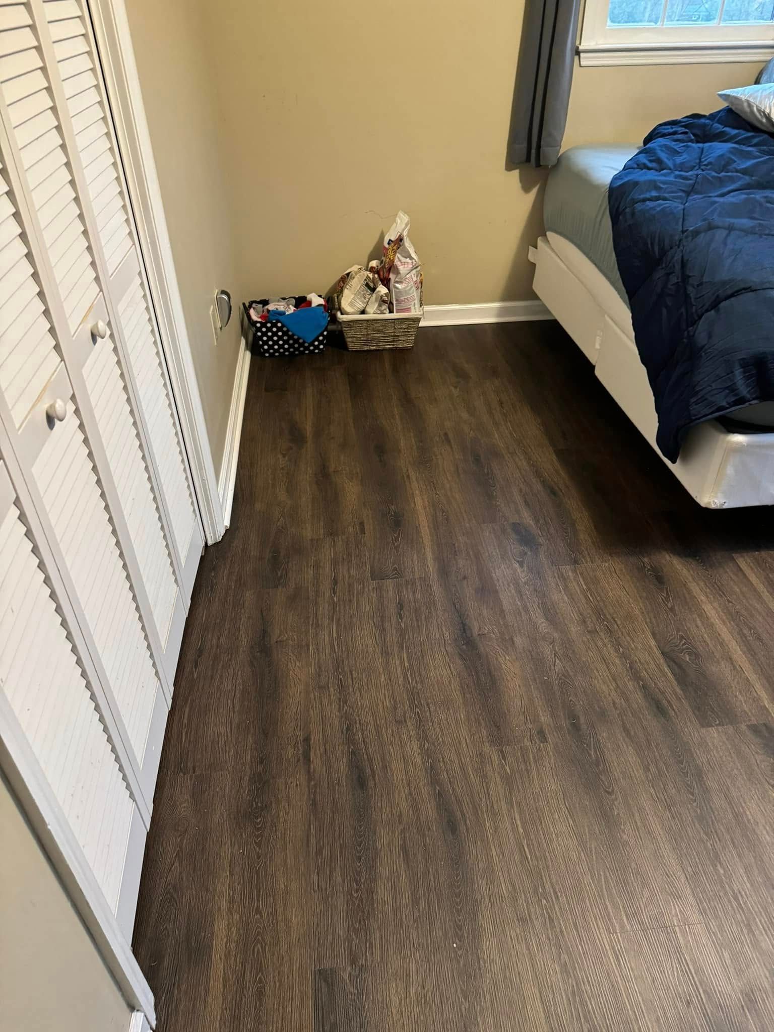 A bedroom with dark wood-look flooring, a white closet, and a bed corner, featuring a basket of items in the corner.