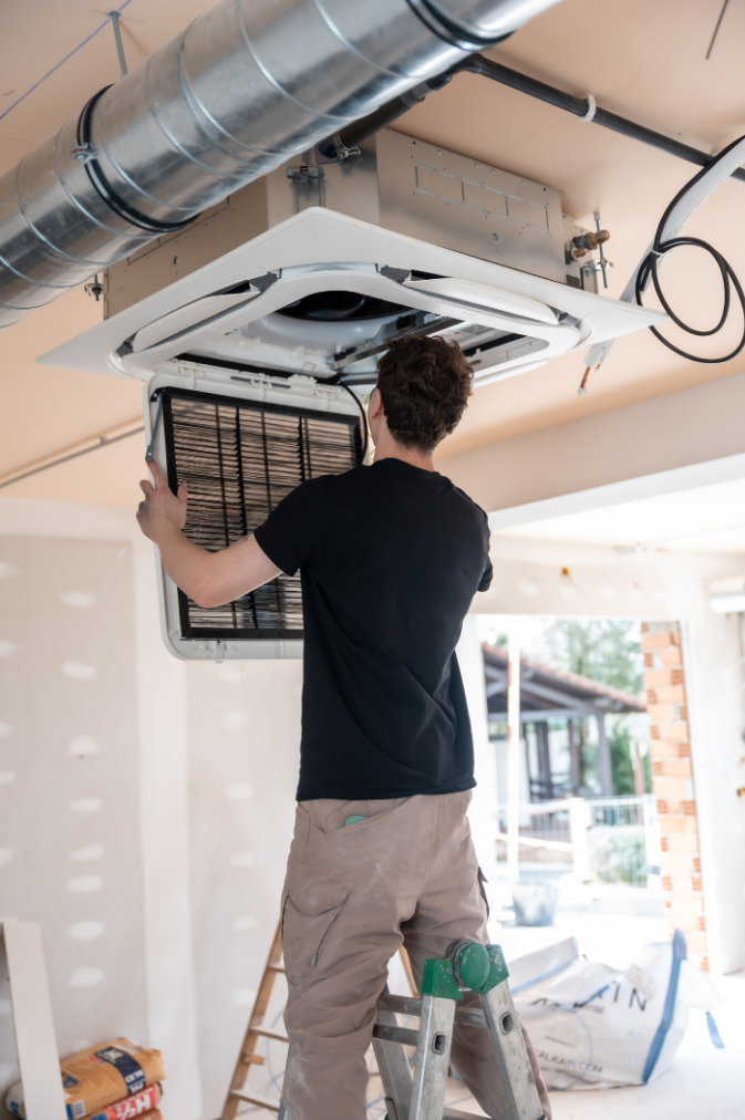 Contact Us A person standing on a ladder while cleaning or installing the filter of a ceiling-mounted HVAC unit.
