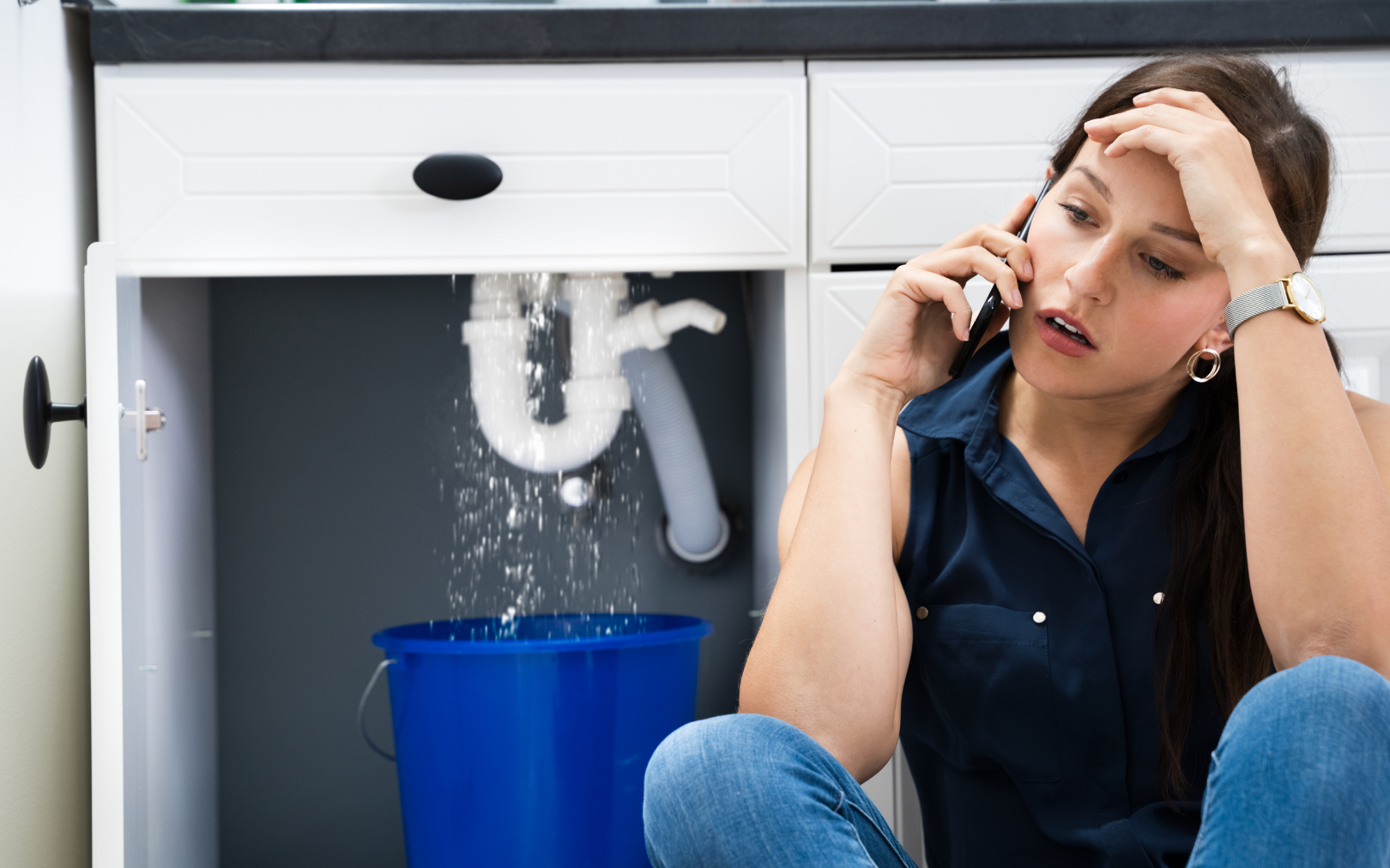 A frustrated person talks on a phone while sitting in front of a kitchen sink with a leaking pipe dripping into a bucket.