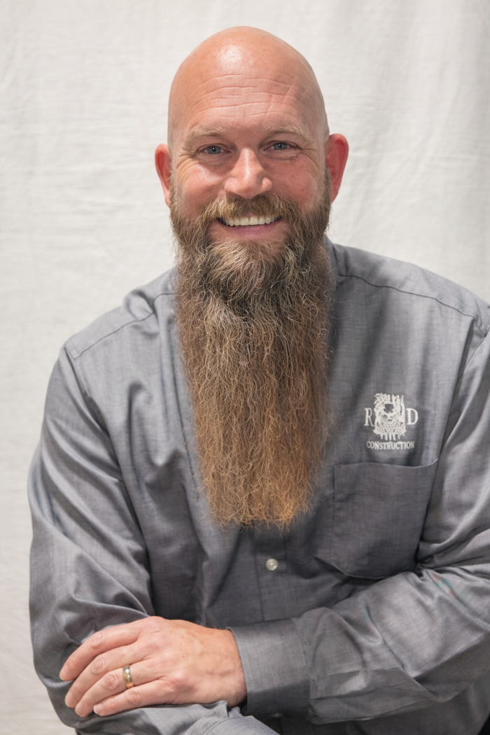 Robert Booher is smiling man with a long beard wearing a gray button-down shirt stands against a plain white background.