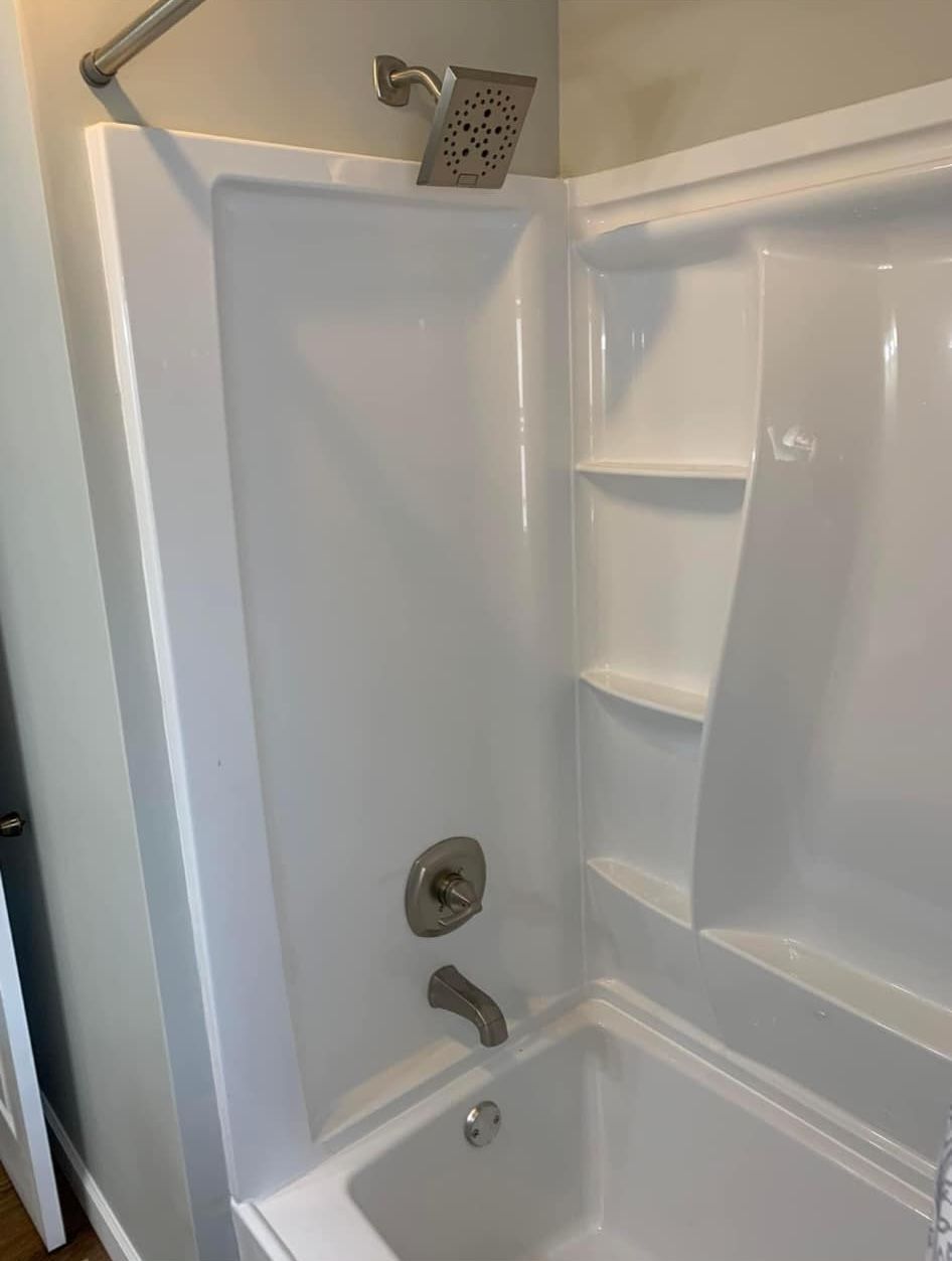 A clean white acrylic bathtub and shower surround with brushed nickel fixtures and built-in storage shelves.