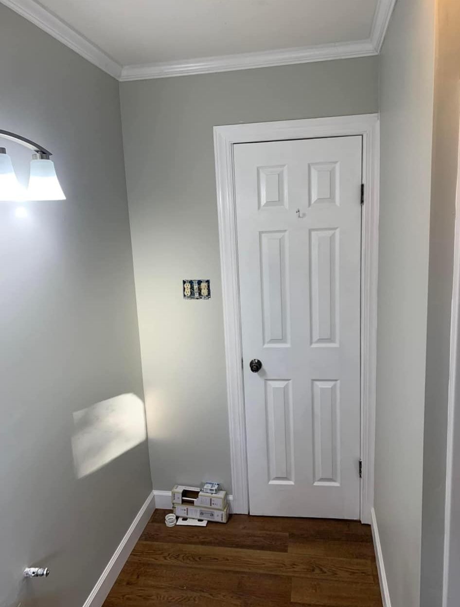 A hallway with gray walls, a white six-panel door, a wall light fixture, and wooden flooring.