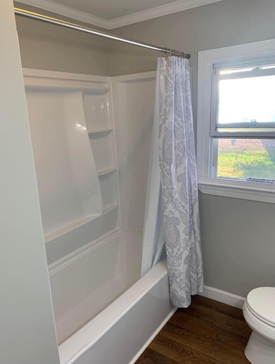 A white bathtub and shower unit with a patterned shower curtain, beside a window and a toilet in a gray bathroom.