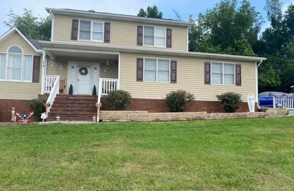 A tan, two-story suburban home with brown shutters, a front porch, and a brick foundation sits behind a mowed green lawn.