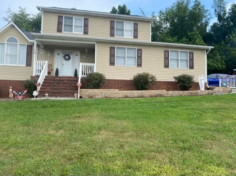 Two-story, tan-sided house with a brick foundation, front porch, and small landscaping bushes in a grassy yard.