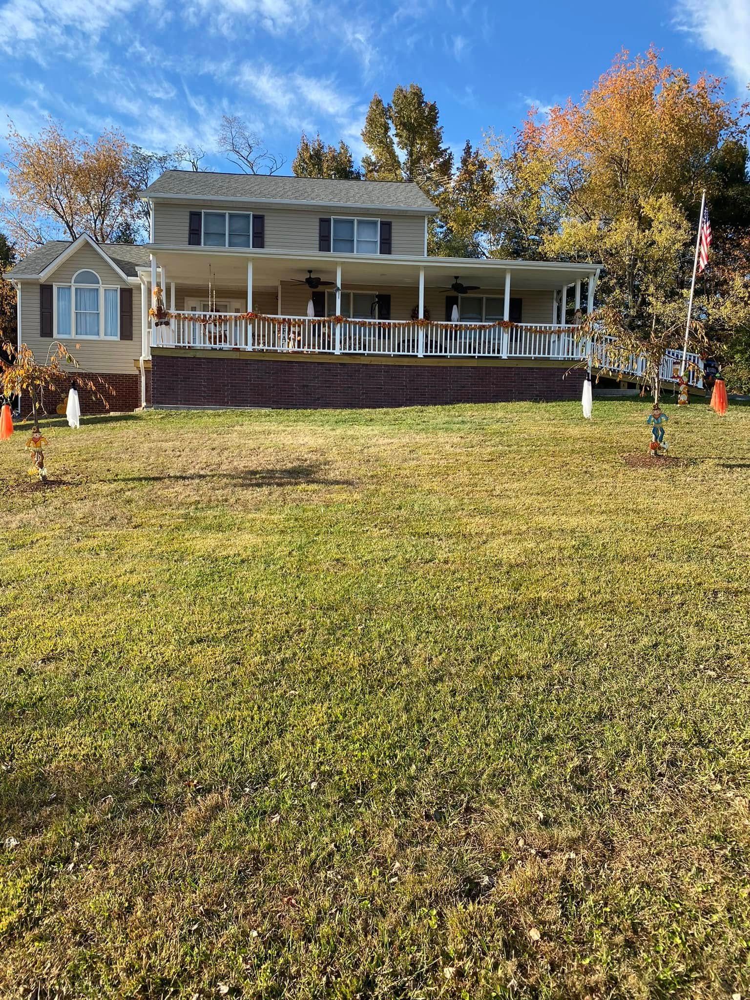 A two-story tan house with a large, covered deck, white railings, and brick foundation, surrounded by trees in autumn.