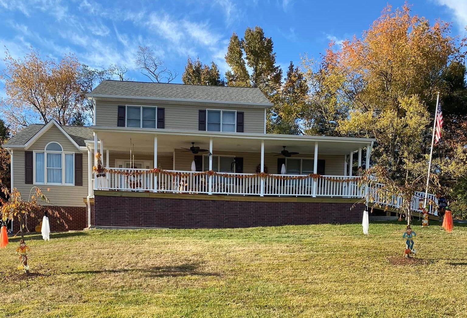 A tan, two-story house with a wide front porch, brick foundation, and autumn trees under a blue sky.