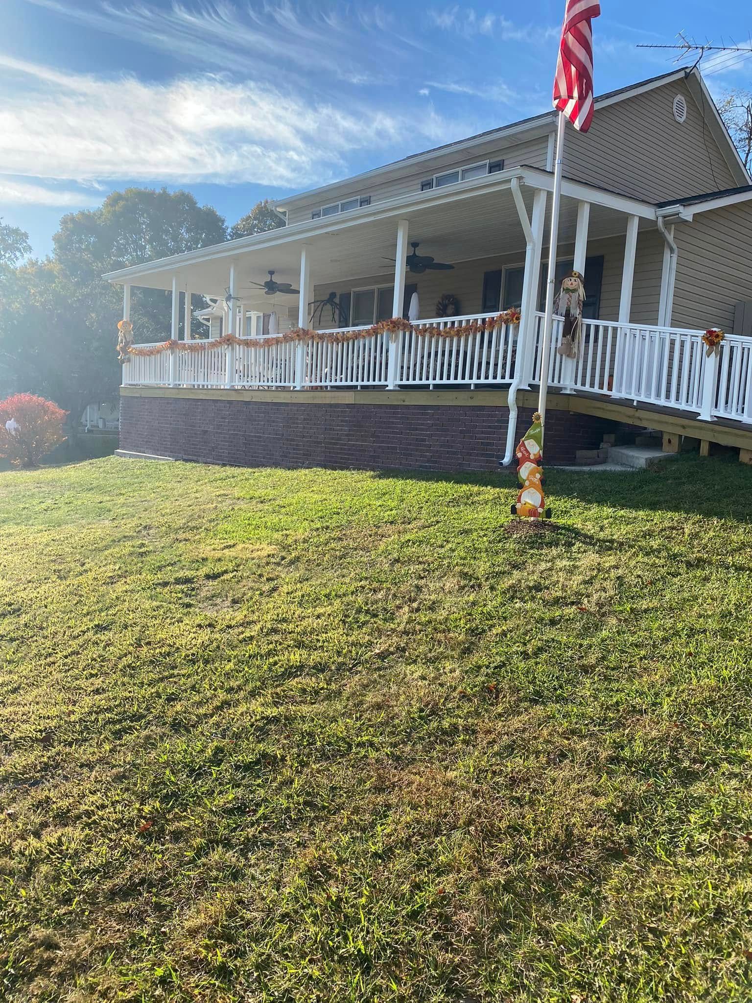A beige, one-story house with a white wrap-around porch, featuring decorative fall garland and a front yard flag.