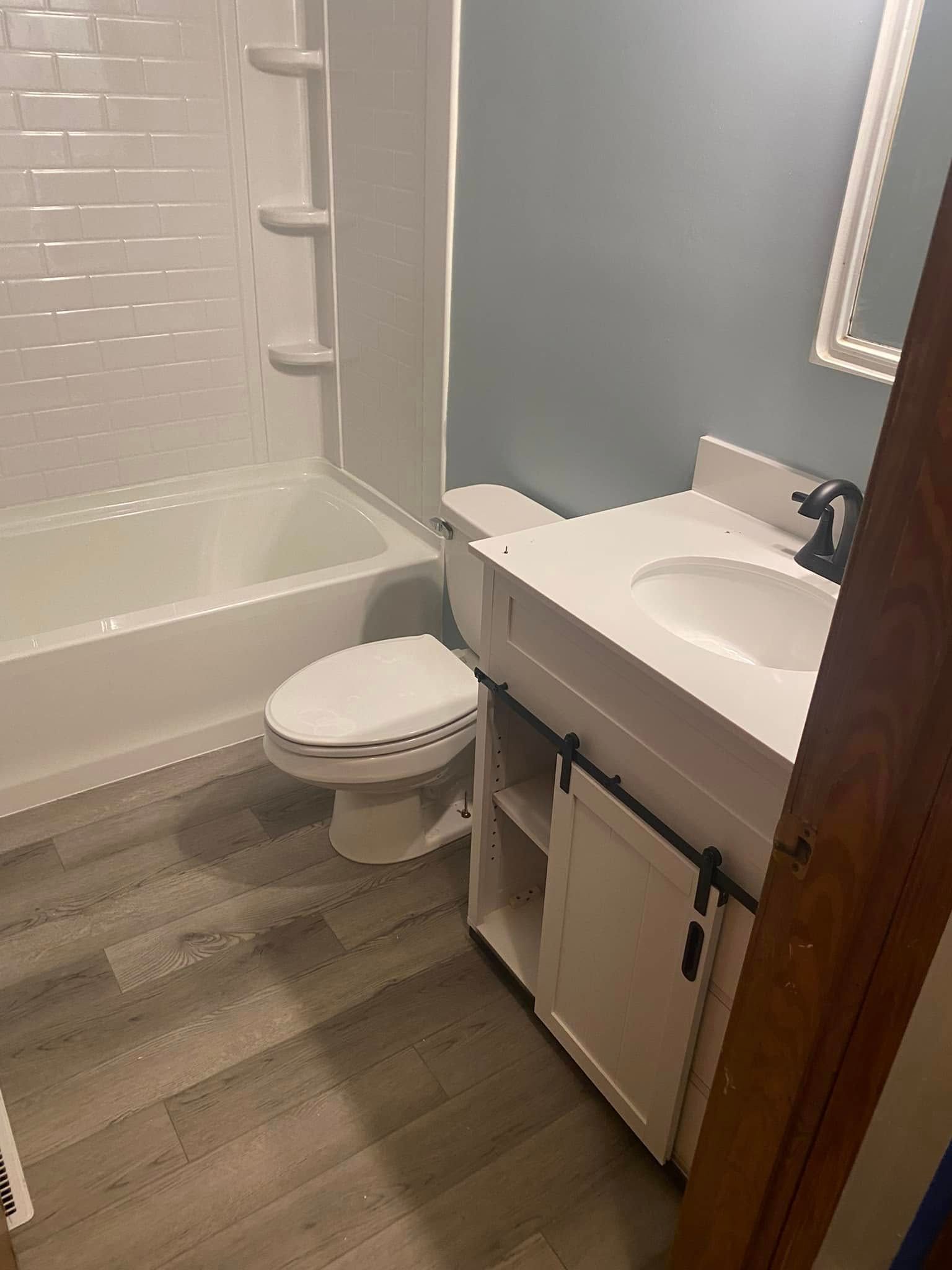 A bathroom featuring a white vanity with a barn-door style cabinet, a toilet, and a bathtub with white tile surround.