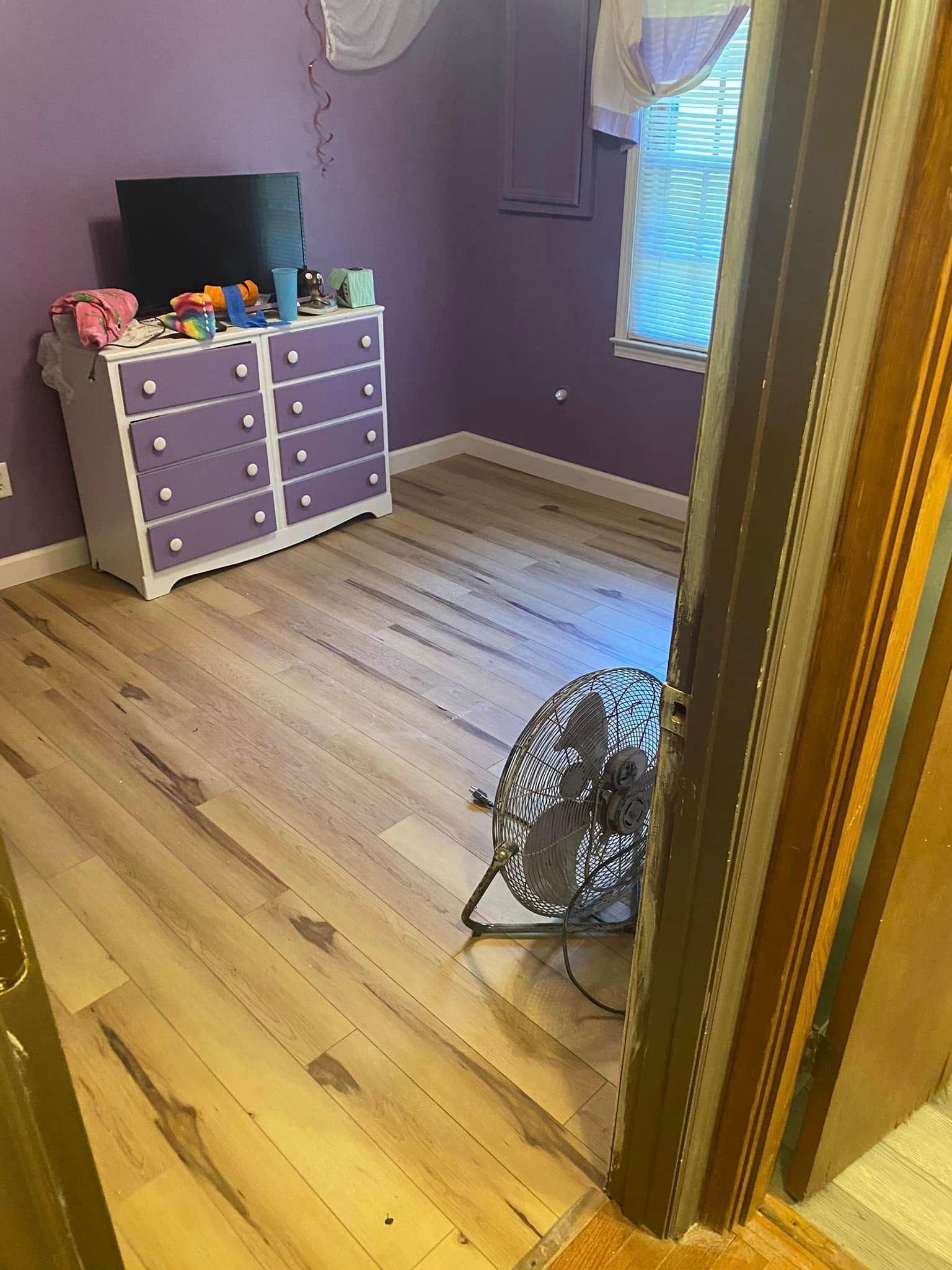 A purple-walled bedroom with a white and purple dresser, a window with curtains, and a metal box fan on the wood floor.