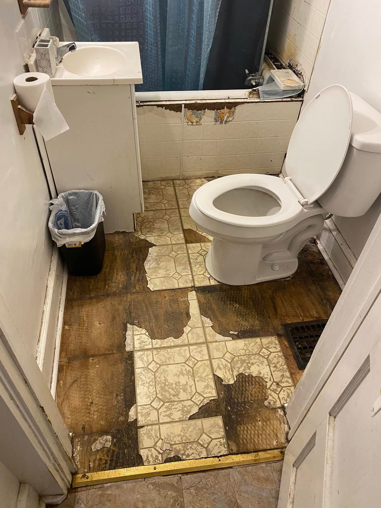 A small, dated bathroom with a white sink, toilet, and bathtub, featuring worn, damaged flooring and peeling wall paint.