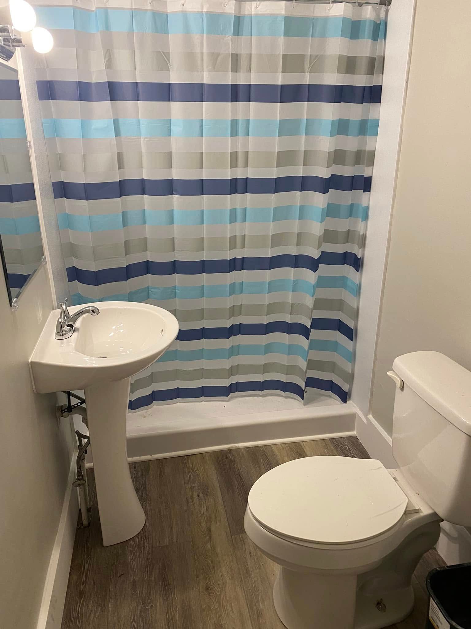 A bathroom featuring a pedestal sink, a toilet, and a shower with a blue, white, and gray striped shower curtain.
